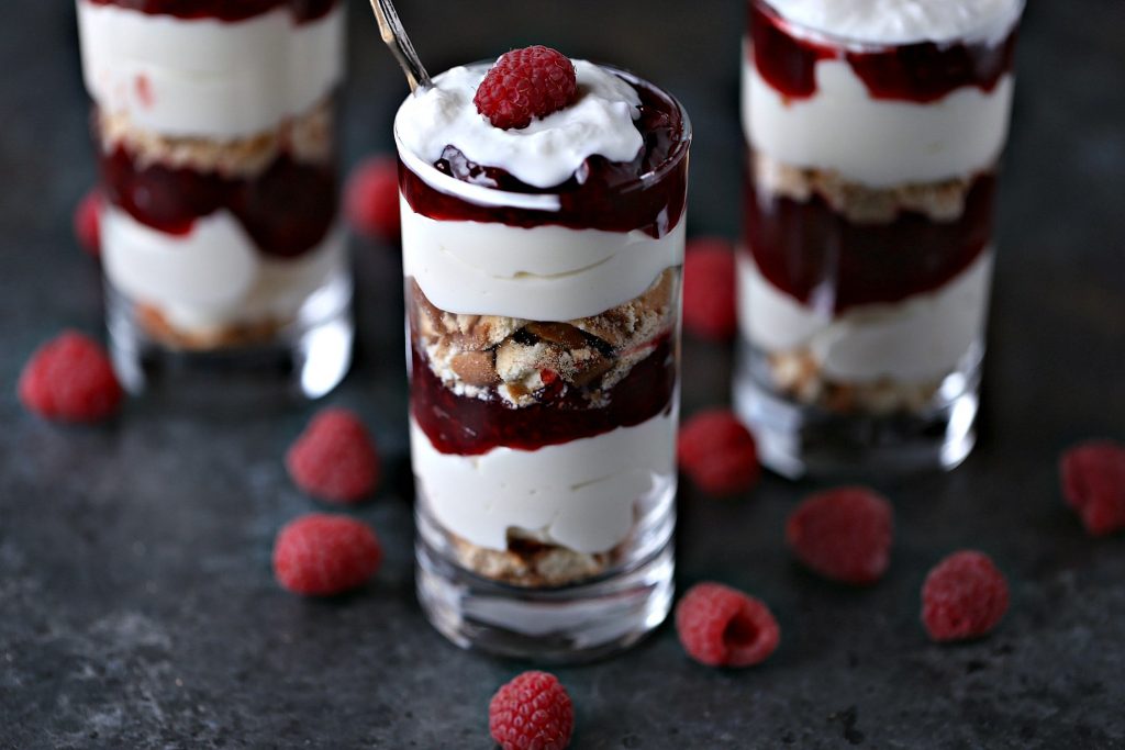 No Bake Lemon Raspberry Cheesecake Parfaits served in mini dessert glasses with raspberries scattered around on a dark surface.