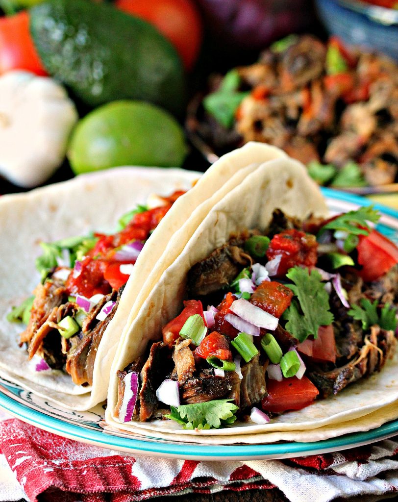 Sweet and Spicy Slow Cooker Carnitas served on tortillas on a plate with ingredients in the background. 