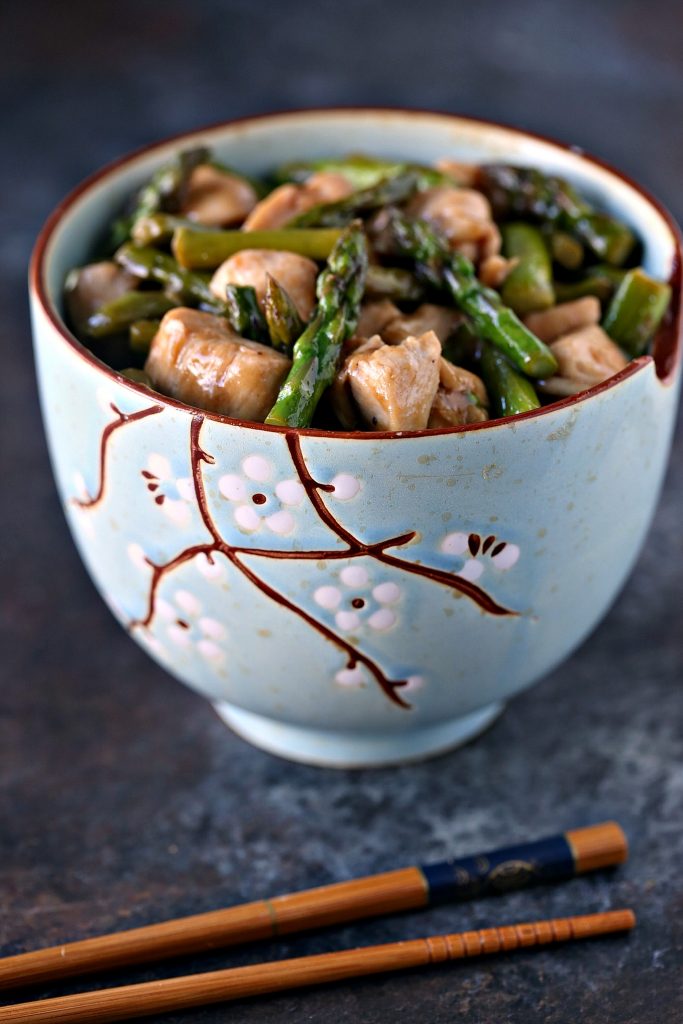 Chicken and asparagus stir fry in a blue oriental style stir fry bowl with wooden chopsticks on counter. 
