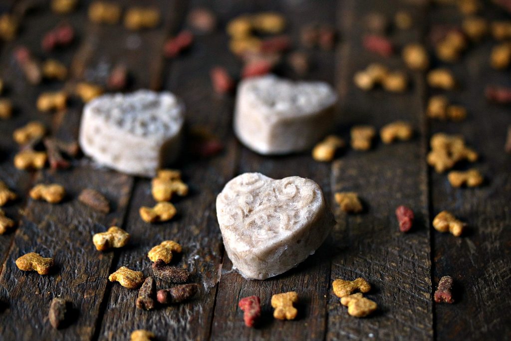 Cat friendly frozen banana bites shaped like hearts on a wood board with tiny cat treats scattered about. 