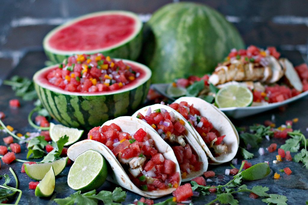 Honey Lime Chicken Tacos with Watermelon Salsa on a dark surface with ingredients scattered around. A hollowed out watermelon filled with watermelon salsa is behind the tacos plus two watermelon halves sliced open. A white plate with sliced chicken, salsa and limes is in the background. 