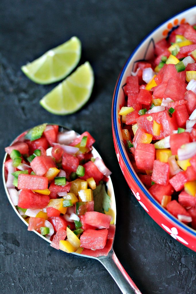 Overhead image of watermelon salsa in a brightly coloured bowl with a spoon laying next to it with salsa inside it, and there are two lime wedges sitting near the spoon and bowl.. Text on image states the recipe name and blog name. 