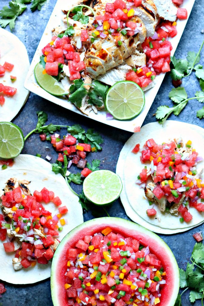 Overhead image of grilled honey lime chicken tacos with watermelon salsa. Watermelon salsa is served in a hollowed out watermelon half. Chicken is on a white plate. Everything is spread out on a greyish blue board.