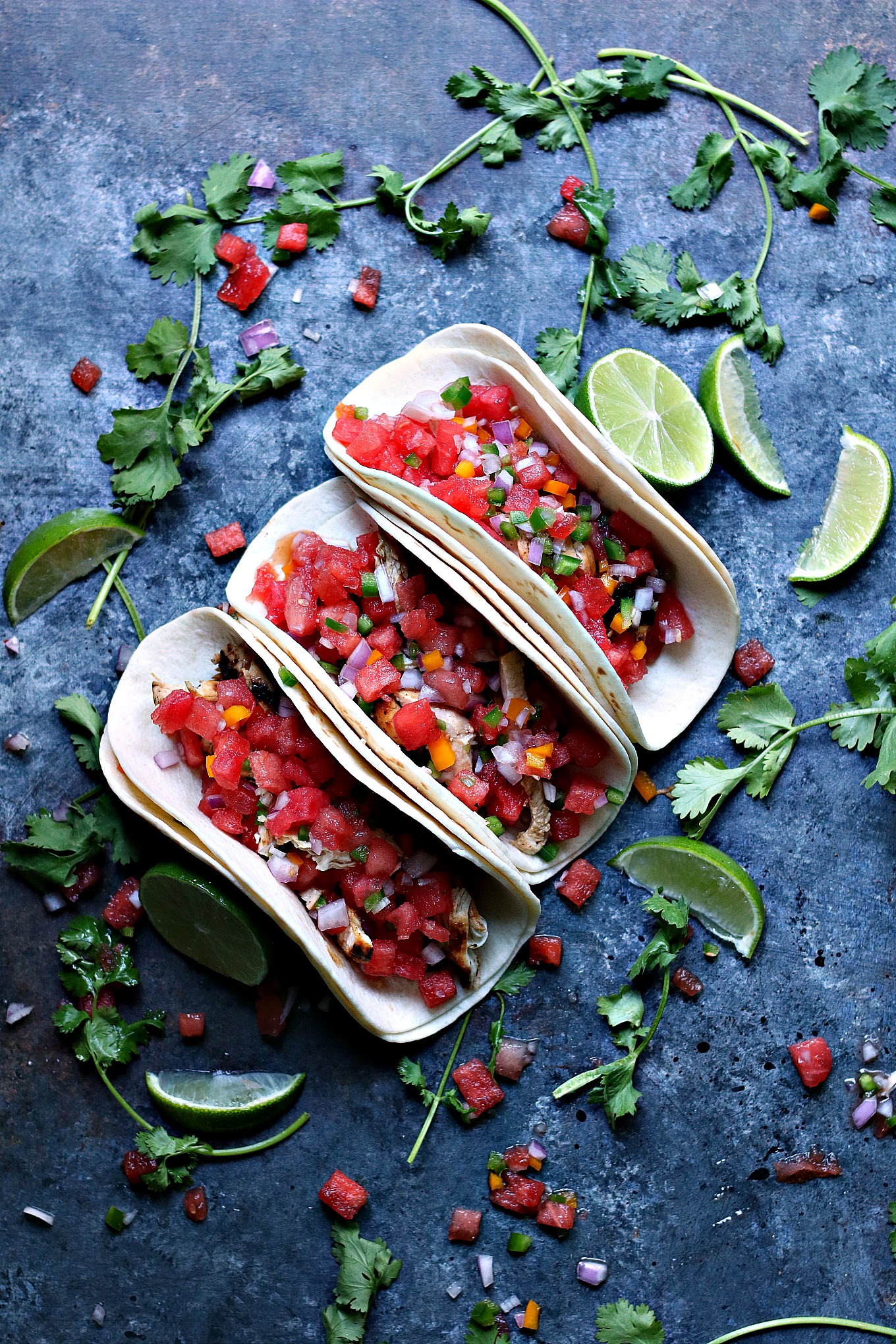 Overhead image of 3 grilled honey lime chicken tacos with watermelon salsa, ingredients are scattered around a dark surface randomly.