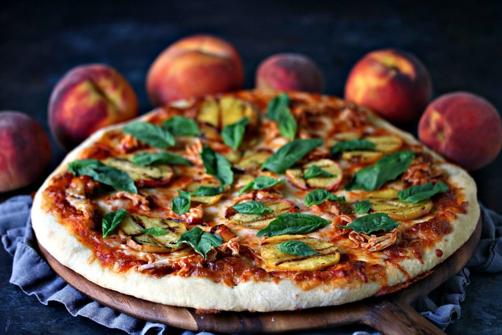 grilled peach and chicken pizza with fresh basil on a wood board with a grey napkin underneath. ripe peaches are in the background and everything is set on a dark counter. 