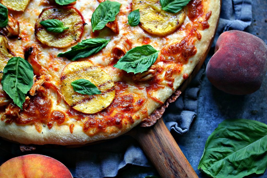 grilled peach and chicken pizza with fresh basil on a wood cutting board with a grey napkin under it, with a dark counter underneath. fresh peaches and basil are scattered about.