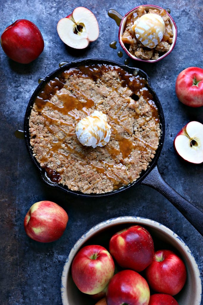 Overhead image of skillet caramel apple crisp that has been baked and is resting on a dark counter. Ice cream tops the apple crisp in the pan and ingredients surround the pan as well. 