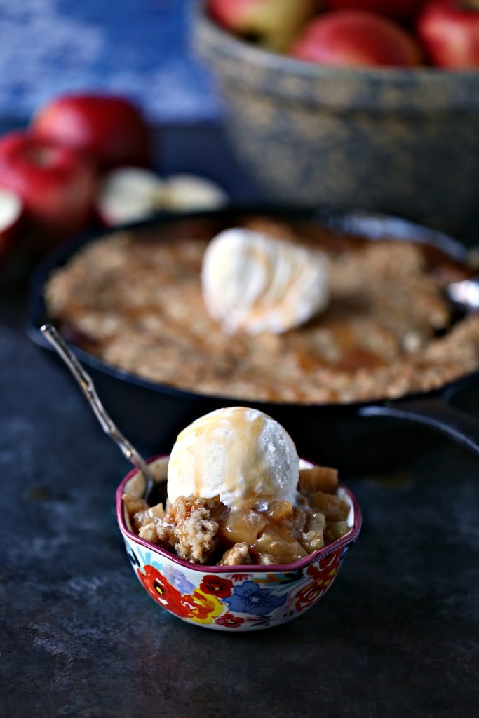 Skillet caramel apple crisp served in a colourful bowl with ice cream on top and a spoon in the bowl. Skillet with remaining apple crisp is in the background and there is also a blue stoneware bowl with apples in it too.