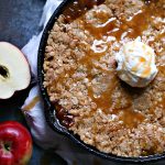 Overhead image of an apple crisp baked in a black cast iron pan that is cooling on a dark counter, ice cream is on top the apple crisp in the middle.