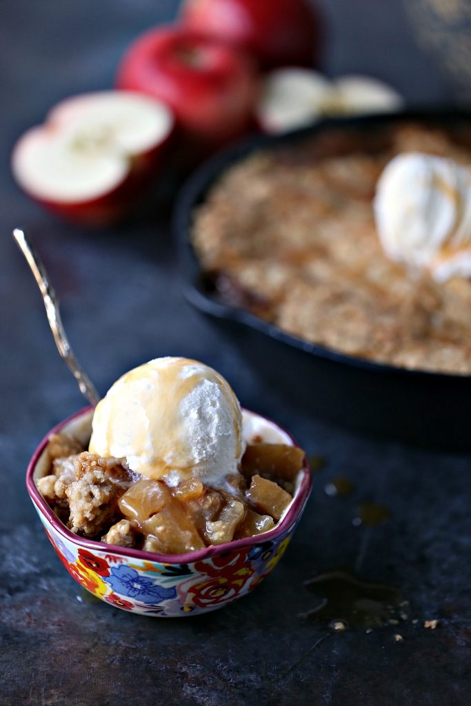 Caramel apple crisp in a colourful bowl with ice cream and a spoon in it. Cast iron pan filled with apple crisp is in the background.