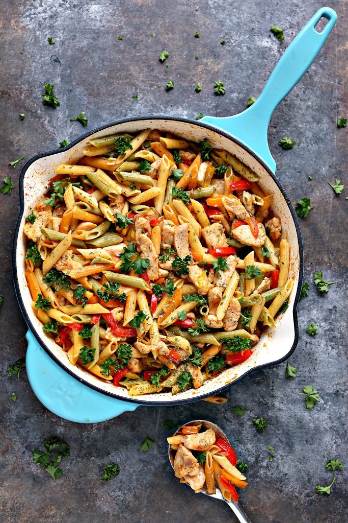 Chicken and vegetable pasta in a blue skillet with a spoonful of food sitting beside it on a counter.