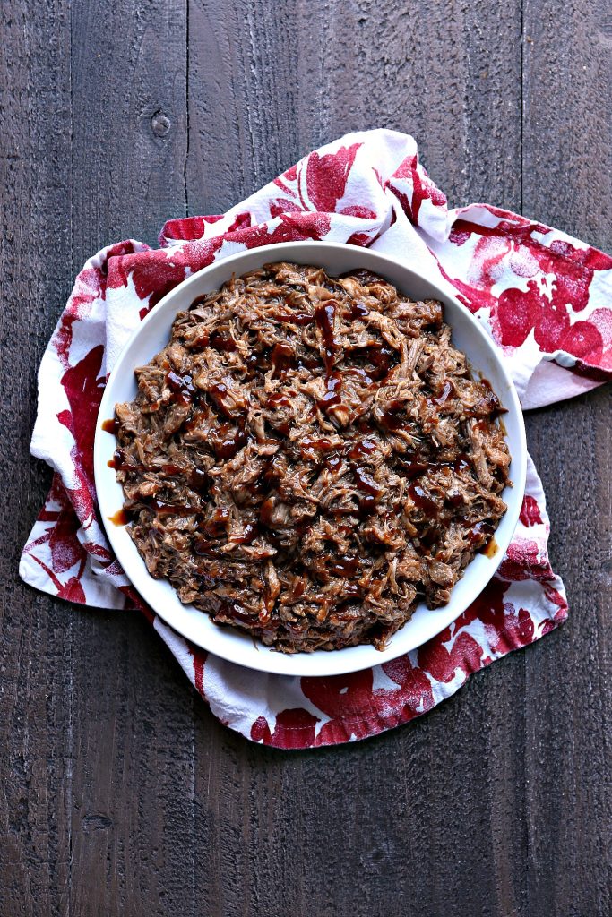 Instant pot pulled pork in a white bowl on top of red and white napkins on a dark wood surface. 