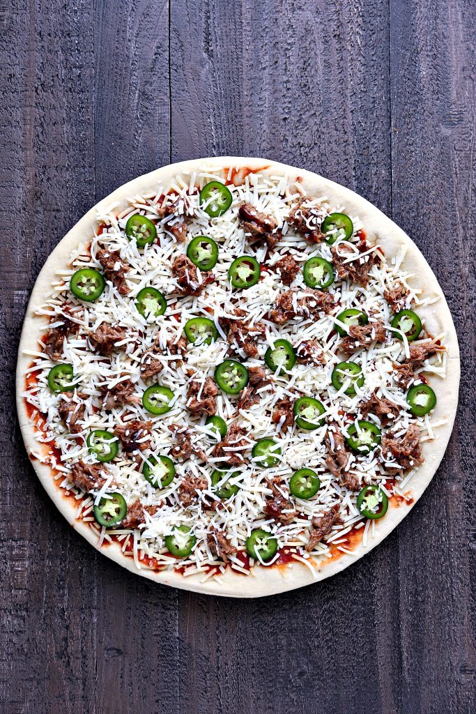 Overhead image of pulled pork pizza topped with peppers, pizza is on a dark wood surface.