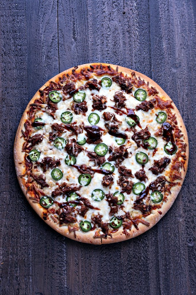 Overhead image of cooked pulled pork pizza with jalapeno peppers on top. Pizza is sitting on a dark wood surface.