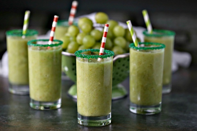 green grape slushies in clear glasses with green sugar rims with a colander of fresh grapes in background