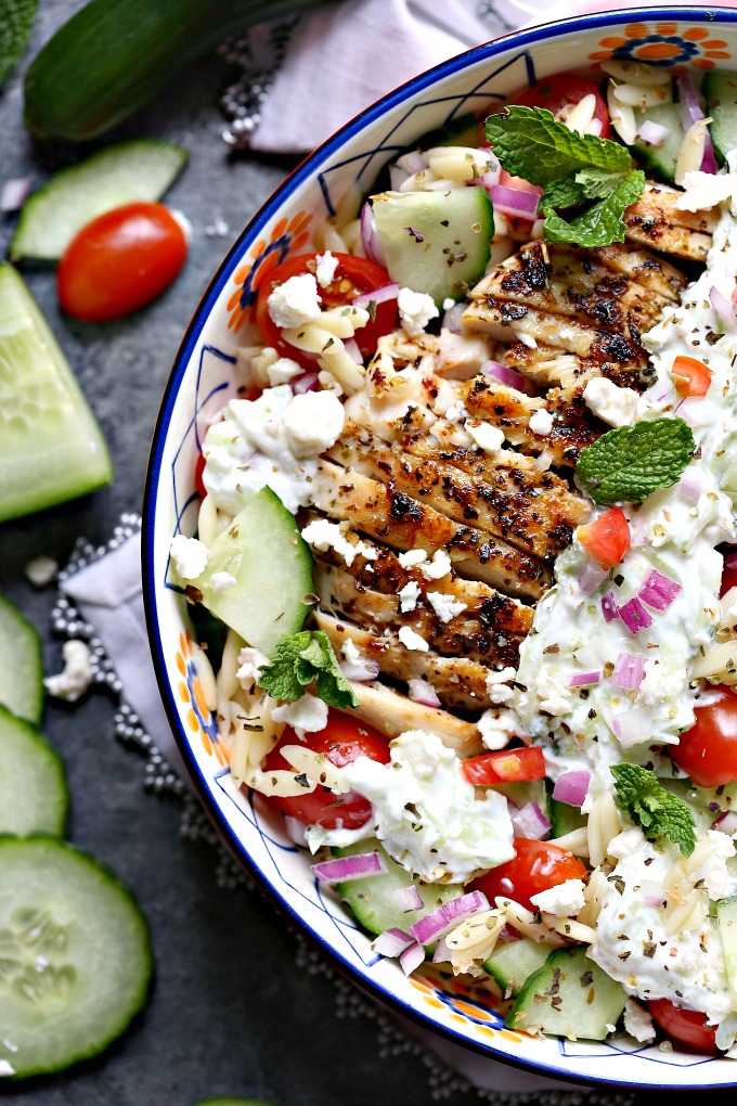 overhead image of Greek chicken orzo power bowls on a white napkin with sliced cucumbers and tomatoes nearby