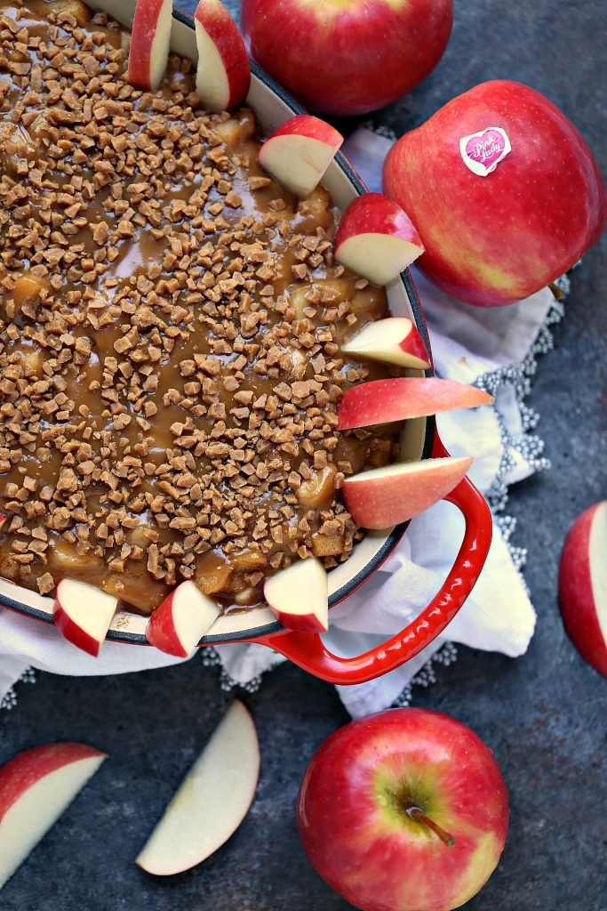 Overhead image of caramel apple dip in a red and white serving dish on a white napkin that is all siting on a dark counter. Whole and cut apples are scattered about and there are some cut apples in the dip ready to be eaten.