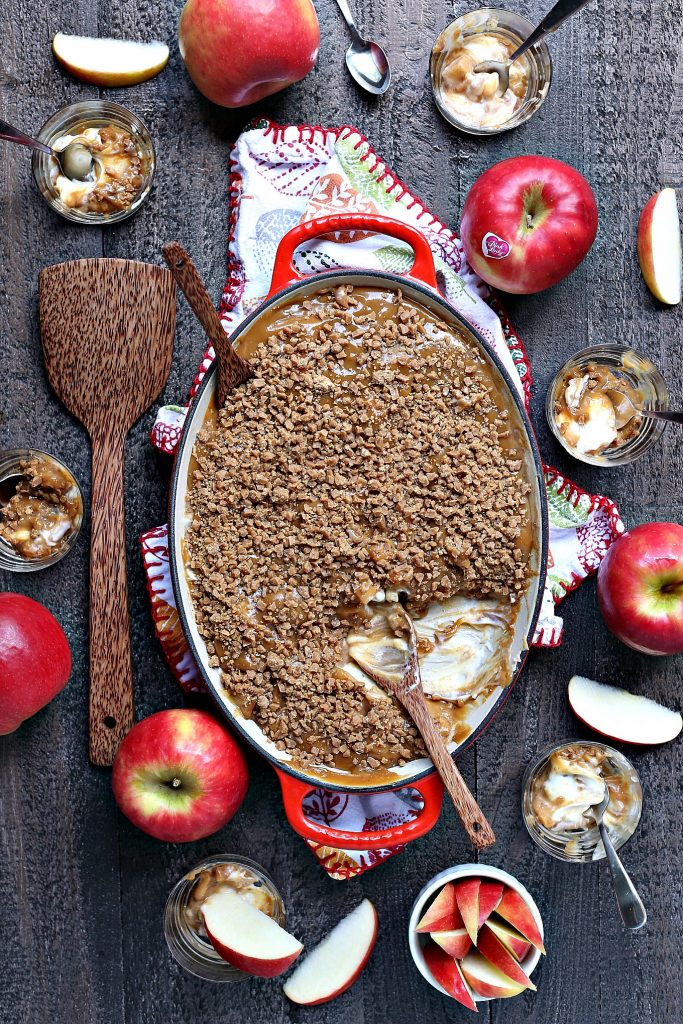 Overhead image of caramel apple dip in a red and white serving dish on a multi-coloured tea towel on a wood surface. Full apples and cut apples are scattered around the serving dish. There's a spoon inside the serving dish and a wooden spatula resting on the wood board. There are also tiny glass dessert dishes with tiny silver spoons.