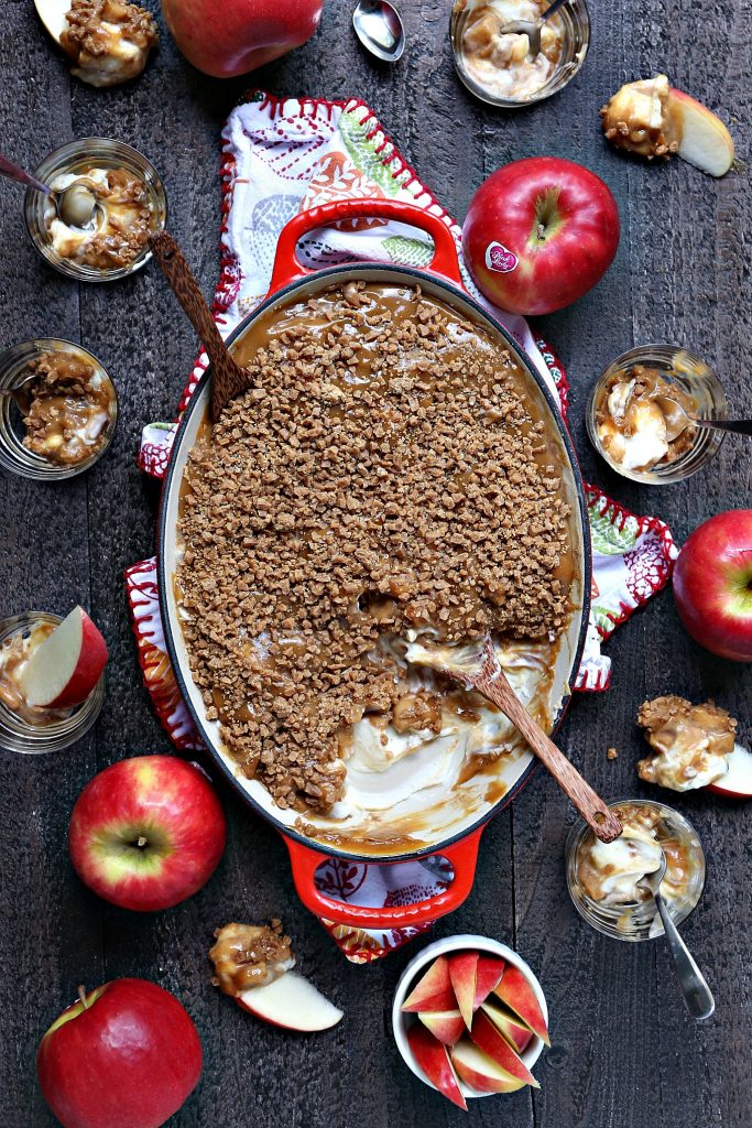 Overhead image of caramel apple dip in a red and white serving dish on a multi-coloured tea towel on a wood surface. Pink lady apples and cut apples are scattered around the serving dish. There's a spoon inside the serving dish and a wooden spatula resting on the wood board. There are also tiny glass dessert dishes with tiny silver spoons.