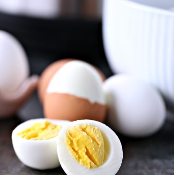 Hard boiled eggs cooked in an instant pot. Image shows an egg cut open in the foreground, behind it is an egg with shell off of half it, with two eggs on either side of it. On the right you can see the corner of an egg in a pink ceramic egg holder. On the right you can see a white bowl and in the middle of the background you see part of the bottom of the instant pot machine.