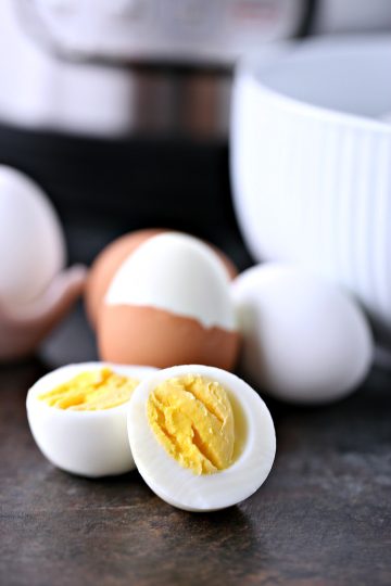 Hard boiled eggs cooked in an instant pot. Image shows an egg cut open in the foreground, behind it is an egg with shell off of half it, with two eggs on either side of it. On the right you can see the corner of an egg in a pink ceramic egg holder. On the right you can see a white bowl and in the middle of the background you see part of the bottom of the instant pot machine.