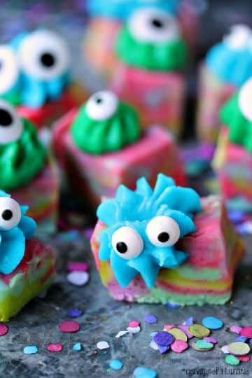 Easy Monster Fudge is brightly coloured swirled fudge cut into squares then topped with icing and googly eyes. Fudge is sitting a grey counter with colourful confetti all around it.