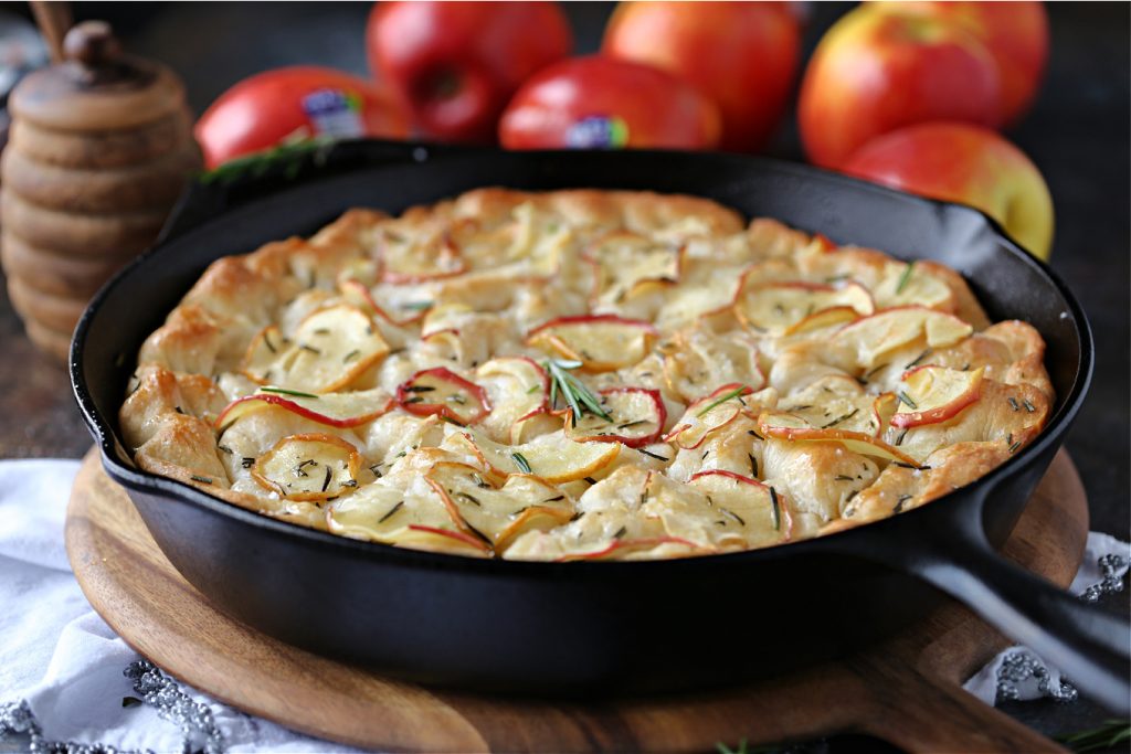 focaccia in a cast iron pan on a wood board with Jazz apples in the background