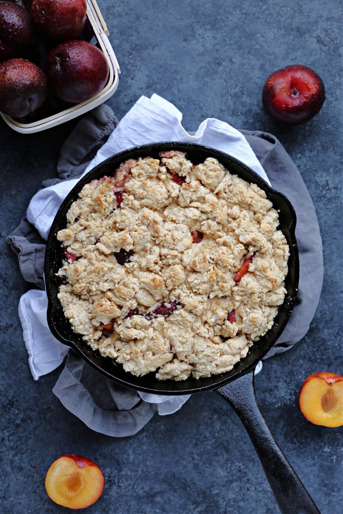 Plum Cobbler in a cast iron skillet with plums scattered on the surface nearby