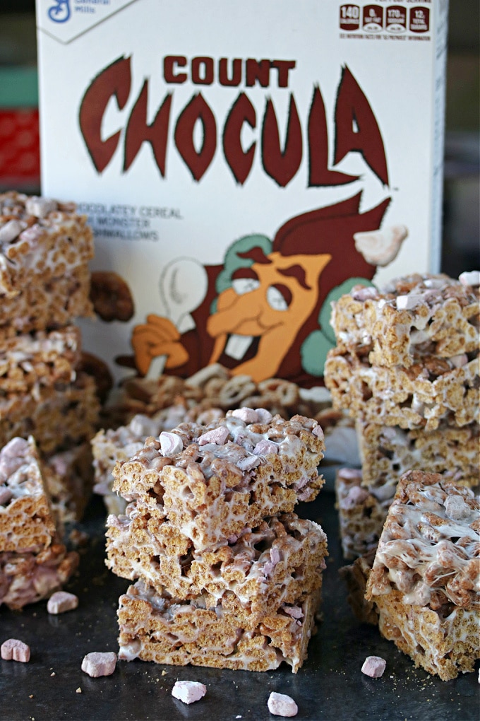 Count Chocula krispie treats stacked on a dark counter with a box of Count Chocula cereal in the background.